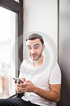 White man sitting on a windowsill in a home environment holding a phone and reading messages at home
