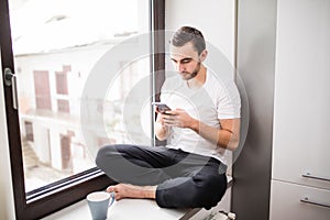 White man sitting on a windowsill in a home environment holding a phone and reading messages at home