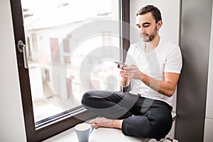 White man sitting on a windowsill in a home environment holding a phone and reading messages at home