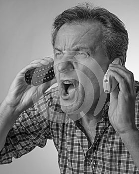 A white man yells while holding two cordless phones, with an expression of great stress, in black and white