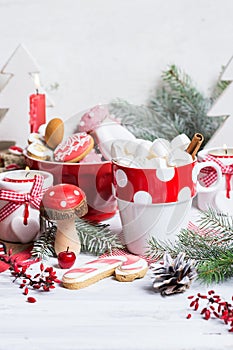 White marshmallows in white red cup with hot chocolate and cinnamon stick on white wooden table decorated with candles, cookies