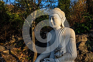 White meditating Buddha statue sits on the rock pile among forest trees in the afternoon time at India. Great religious