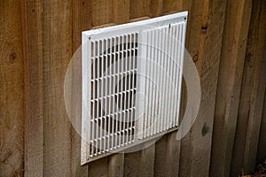 A white metal air vent grille on the side of a wooden building