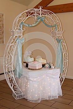 White metal arch over a pastle wedding cakes.bows and flowers