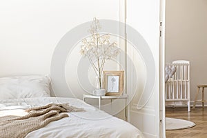 White metal bedside table with coffee mug, twig in glass vase and simple poster in frame placed by the bed