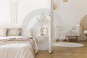 White metal bedside table with decor and coffee mug placed by the bed in bright bedroom interior with door to newborn baby room