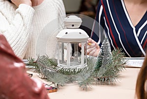 A white metal candlestick with candle inside surrounded with christmas tree. Women sitting around the table. Christmas party or