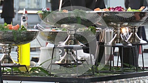 White metal dishes with food are on the tables, the party is outdoors, waiters serve customers.