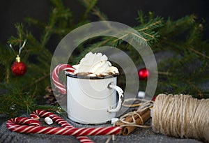 White metal mug with hot drink with whipped cream and Lollipop on the background of fir branches with red balls, coil of rope,