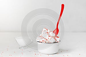 White milk ice cream with red and pink sprinkles decoration and a red plastic spoon in a white metal mug on a white background