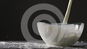 White milk is poured into a glass bowl on a black background. The milk splash falls out of the bowl on the gray table