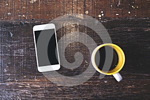 A white mobile phone with blank black desktop screen with coffee cup on vintage wooden table