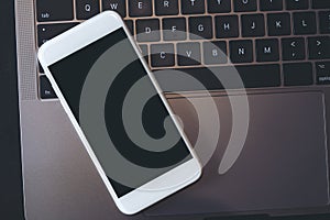 A white mobile phone with blank black desktop screen on a computer laptop keyboard