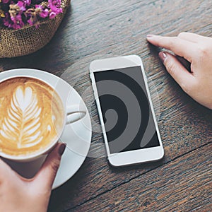 White mobile phone with blank black screen and hand holding hot latte coffee on vintage wood table in cafe