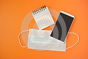 White mobile phone with clipping path on touch screen, medical mask and white opened calendar on vivid orange background