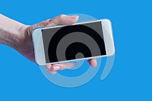 White mobile phone in left female`s hand on blue background with copy space