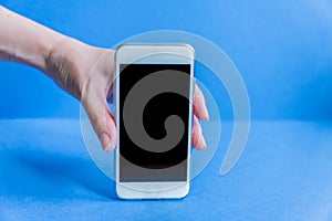 White mobile phone in left female`s hand on blue background with copy space