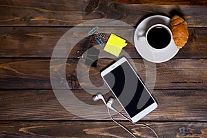 White mobile phone on wooden wooden background with headphones, cup of coffee, croissant and stationery appliances