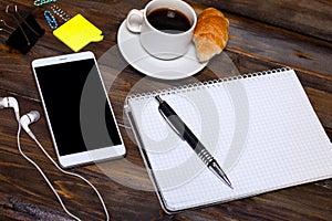 White mobile phone on wooden wooden background with headphones, cup of coffee, croissant and Stationery appliances