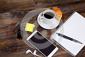 White mobile phone on wooden wooden background with headphones, cup of coffee, croissant and Stationery appliances