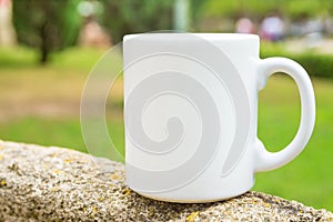 White mockup coffee or tea mug on standing on stone outdoors. Nature background with green trees grass. Summer spring. Blank space