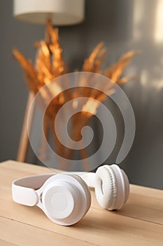 White modern wireless headphones on wooden table and bouquet of dry flowers over gray house wall with sunlight