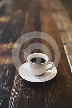 A white mug with black coffee and a saucer stand on a wooden table.