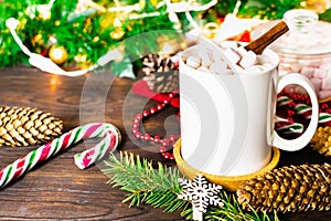 White mug of cocoa with marshmallows, lollipops, fir cones, Christmas tree branch, garland and snowflake on wooden table