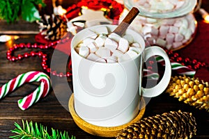 White mug of cocoa with marshmallows, lollipops, fir cones, Christmas tree branch, garland and snowflake on wooden table