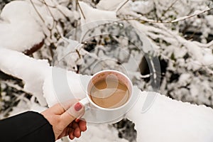 White mug of coffee with milk in the girl`s hand on the snowy background, winter concept photo.