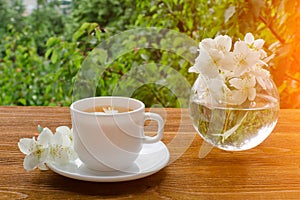 White mug of tea and a vase with jasmine on a wooden table, greens on the background