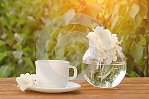 White mug of tea and a vase with jasmine on a wooden table, greens on the background, sunlight