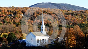 A white new england church with a hillside covered in fall foliage at stowe in vermont