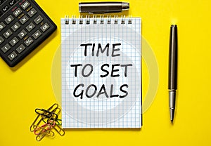 White note with inscription `time to set goals` on beautiful yellow background, colored paper clips, metalic pen, cap and