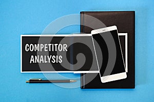 A white notebook with black pages, a smartphone and a pen on a blue background. The inscription COMPETITOR ANALYSIS