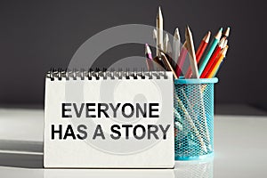 A white notebook with the inscription EVERYONE HAS A STORY standing near a blue pencil holder on white and gray background.