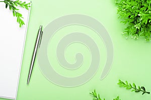 White notebook with steel pen and green branches on a green background. Office table, spring composition, copy space