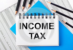 White notebook with the text INCOME TAX on the table next to black pencils on a blue background and reports. Business concept