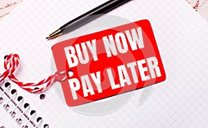 On a white notebook there is a black pen and a red price tag on a string with the text BUY NOW PAY LATER