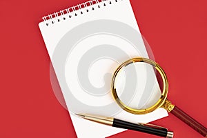 White notepad with a stylish pen, magnifying glass on a red background. Copy space