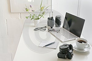 White office desk with laptop, camera, lenses, cable, glasses, notepad and a coffee cup, modern workplace of a photographer