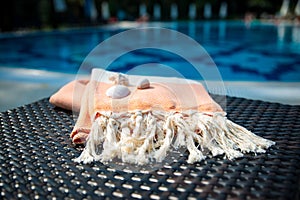 A white and orange Turkish peshtemal / towel and white seashells on rattan lounger with blue a swimming pool as background.