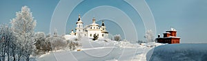 White orthodox church amidst deep snow and bare trees on the background of clear blue sky panorama