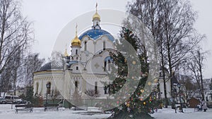A white Orthodox church with gilded domes and crosses is surrounded by trees.Next to it stands a large Christmas tree, decorated w