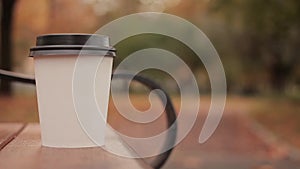 White paper cup with hot drink in autumn city park close up with defocused background skater boy walking people