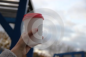 white paper cup with red plastic lid in the woman& x27;s hand against the cloud sky, place for text