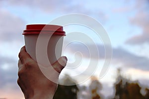 white paper cup with red plastic lid in the woman& x27;s hand against the cloud sky, a place for text on the right
