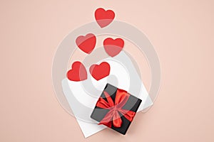 White paper envelope and Valentines hearts on pink table. Black gift box with red ribbon on festive background.