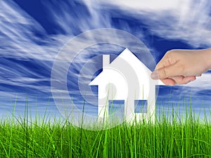 White paper house held in hand by a man in a green summer grass over a blue sky background with clouds