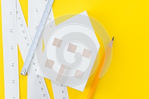 White paper house, yellow pencil and white measuring tapes with centimetre and inches on vivid yellow background, architecture,